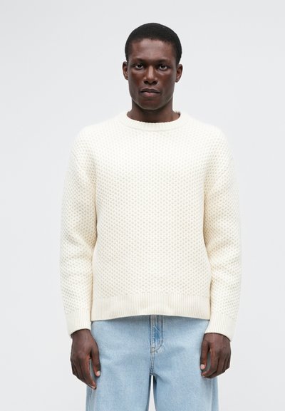 Pulover crem din tricot gros cu guler rotund și mansete cu nervuri. Asortat cu blugi de denim de un albastru deschis. Textura este proeminentă.