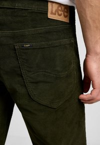Pantaloni in corduroy verde oliva con superficie testurizzata, caratterizzati da una toppa in pelle con il marchio "Lee" sulla vita e tasche posteriori cucite.