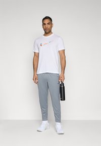Hvit Nike T-skjorte med et flerfarget logo, grå treningsbukser og hvite joggesko. Holder en svart vannflaske.