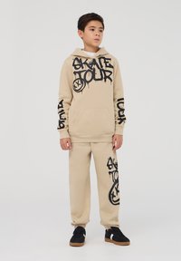 Beżowy hoodie z czarnym tekstem w stylu graffiti oraz dopasowanymi spodniami dresowymi. Materiał wydaje się miękki; posiada kieszeń z przodu i elastyczne mankiety.