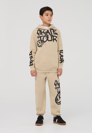 Felpa beige con testo in stile graffiti nero e pantaloni abbinati. Il materiale sembra morbido; presenta una tasca frontale e polsini elasticizzati.
