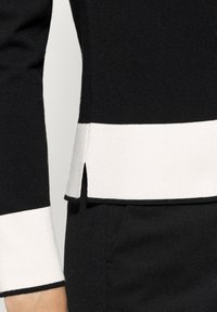 Gestreiftes Strickoberteil in Schwarz und Weiß mit enger Silhouette, das einen gespaltenen Saum und strukturiertes Material aufweist. Die Manschetten haben ein kontrastierendes weißes Band.