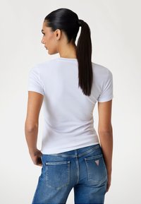 T-shirt bianco a maniche corte visto da dietro, abbinato a jeans in denim blu con un logo triangolare sulla tasca posteriore destra.