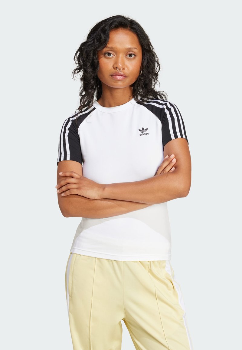 Baltas sportinis marškinėliai su juodomis raglan apykaklėmis ir trimis vertikaliais juodais dryžiais ant kiekvieno peties, su juodu Adidas logotipu ant krūtinės.