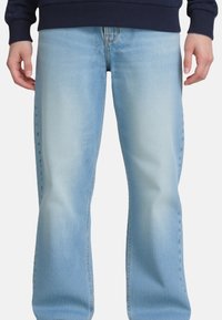 Persona che indossa jeans azzurri sbiaditi e una maglia a maniche lunghe blu navy, in piedi con le mani rilassate ai lati su uno sfondo semplice.