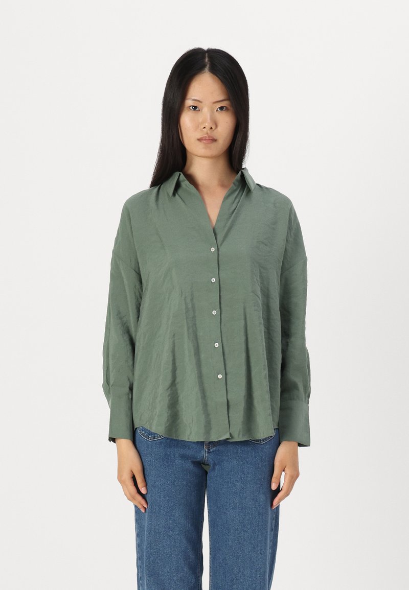 Grønn skjorte med avslappet passform, som har spiss krage, lange ermer og glatt tekstur. Kombinert med blå denimjeans.