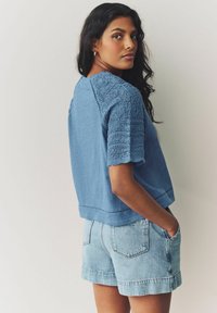 Blue top with textured, embroidered short sleeves; paired with light denim shorts. Fabric appears soft with a relaxed fit and simple design.