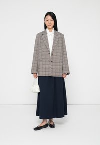 Overmaatse geruite blazer in grijs en bruin, witte blouse, marineblauwe geplooide rok, zwarte flats en kleine witte gestructureerde handtas.
