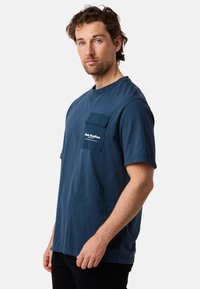 Meriharmaa puuvillapaita, jossa on pyöreä kaula-aukko, lyhyet hihat ja vasemmassa rinnassa läpällä varustettu tasku, jossa on painettu logo.