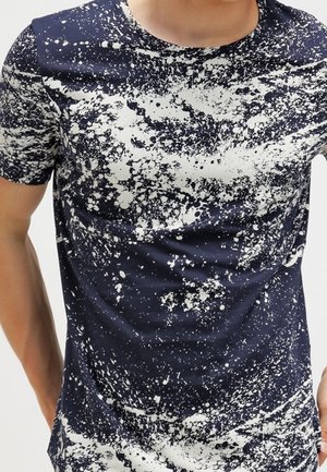 Camisa de manga corta azul marino con un patrón abstracto de salpicaduras de pintura blanca, usada por una persona con los brazos ligeramente doblados.