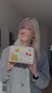 Kreemivärvi Coachi kotike, millel on tikitud kujundused, sealhulgas kirsid, liblikas, NYC ja pretsel, ning hõbedane tõmblukk.