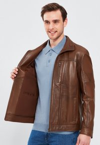 Giacca in pelle marrone con chiusura a zip, dotata di tasche laterali e tasca sul petto, texture liscia e colletto alto, indossata sopra una polo blu.