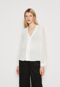Camicia bianca e trasparente con scollatura a V e maniche lunghe. Presenta una chiusura con bottoni e un orlo dritto. Abbinata a pantaloni larghi neri con una texture.