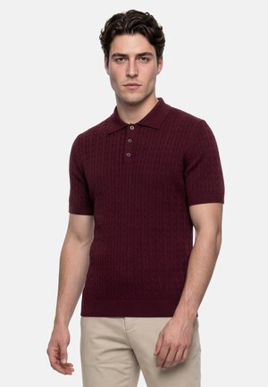 Junger Mann trägt ein burgunderfarbenes, kurzärmeliges, zopfgestricktes Polohemd und beigen Hosen, steht vor hellem Hintergrund.