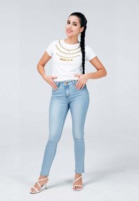 T-shirt bianco con dettagli in perline dorate, abbinato a jeans skinny azzurri e tacchi bianchi con cinturini. Texture liscia, design aderente.