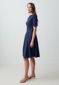 Donkerblauwe jurk tot de knie met korte mouwen, aansluitend lijfje en uitlopende rok. Gemaakt van textuurstof met een strak ontwerp.