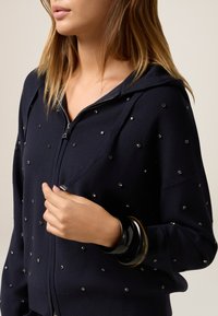 Veste à capuche zippée bleue marine avec un tricot texturé, ornée de petits clous métalliques. Elle possède une capuche et des poignets côtelés. Des bracelets noirs et écaille de tortue sont portés.