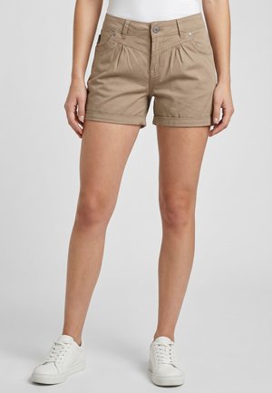 Woman wearing beige cuffed shorts paired with white sneakers, standing against a plain light background.