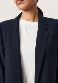 Námořnický blazer se strukturovanými klopami, vyrobený z měkk�ého materiálu, vrstvený přes bílý top s hladkou texturou a kulatým výstřihem.