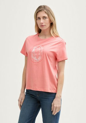 Koralen bombažni T-shirt z ohlapnim krojem, kratkimi rokavi in krožno belo grafiko na sredini. V kombinaciji z modrimi jeans hlačami.