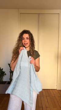 Νεαρή γυναίκα με σγουρά μαλλιά κρατάει ένα ανοιχτό μπλε πλεκτό πουλόβερ εσωτερικού χώρου, στέκεται σε ξύλινο πάτωμα κοντά σε μπεζ τοίχο και φυτά.