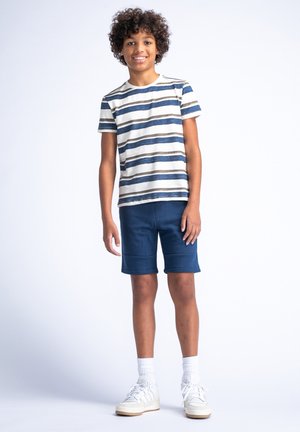 Gestreept t-shirt in crème, blauw en bruin; marineblauwe shorts; witte sokken; witte sneakers. Casual outfit geschikt voor warm weer.