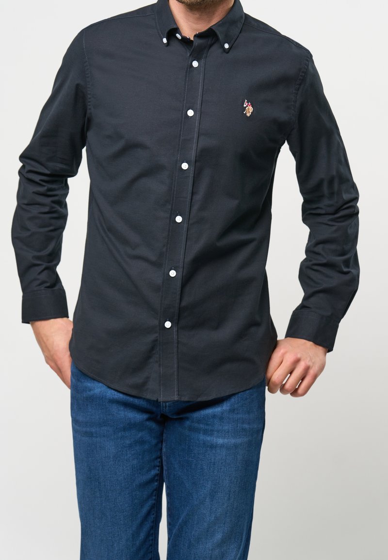 Chemise bleu marine à col pointu, avec des boutons blancs et un petit détail brodé sur la poitrine, associée à un jean bleu.