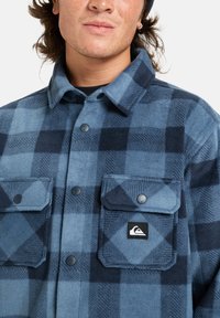 Camisa de franela a cuadrícula en azul y marino con dos bolsillos en el pecho y botones de presión negros. Presenta un logo de la marca tejido en el bolsillo izquierdo.