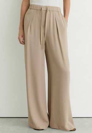 Pantaloni beige a gamba larga con vita elastica e coulisse, indossati con le mani nelle tasche e top bianco visibile.