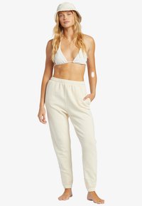 Pantalones deportivos de color claro con cinturilla y bolsillos laterales, combinados con un top de bikini blanco a rayas y un sombrero de cubo claro.