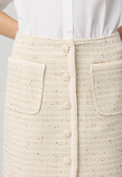 Jupe beige texturée avec des accents de fil doré, dotée de six boutons et de deux poches plaquées. Fabriquée en tissu tissé avec un design structuré.