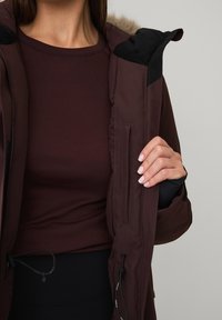 Bruine geïsoleerde jas met een met bont gevoerde capuchon, met rits afsluitbare zakken en trekkoorddetails. Daaronder een nauwsluitend, donkerbordeaux rood shirt met lange mouwen.