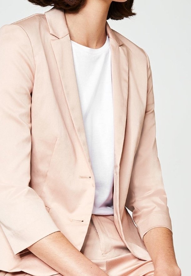 Femme portant un blazer rose clair et un pantalon assorti avec un t-shirt blanc à col rond, assise les mains posées sur ses genoux.