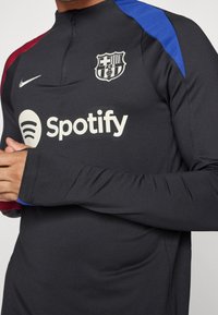 Schwarzes Nike Trainingstop mit roten und blauen Schulterakzenten, Barcelona-Logo und Spotify-Logo auf der Vorderseite. Design mit hohem Kragen.