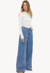 Mujer joven con un top blanco ajustado de manga larga y vaqueros anchos azules decorados con tachuelas de colores, de pie con una mano en el bolsillo.