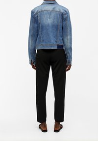 Giacca di jeans in blu chiaro, design aderente, con due tasche posteriori e polsini con bottoni; abbinata a pantaloni neri e sandali.