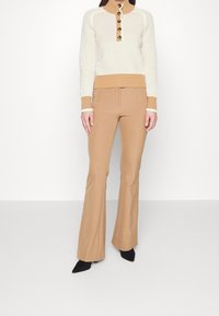 Kobieta w beżowych spodni flared i białym swetrze dzianinowym z jasnobeżowym kołnierzem i mankietami, w czarnych spiczastych butach, stojąca przy białej ścianie.