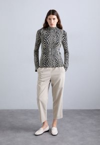 Zwart-wit gestreepte lange mouwtop met een hoge kraag, gecombineerd met lichtbeige broek en witte loafers op een grijzige achtergrond.