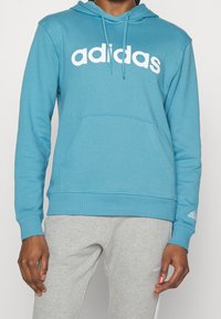 Blå hoodie i mjukt tyg. Har en vit "adidas"-logotyp över bröstet, ficka fram och ribbade ärmar. Matchas med grå sweatpants.