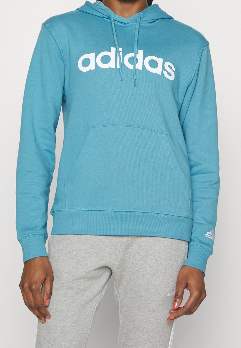 Blå hoodie i mjukt tyg. Har en vit "adidas"-logotyp över bröstet, ficka fram och ribbade ärmar. Matchas med grå sweatpants.