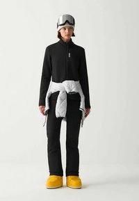 Completo da sci con giacca nera con zip, pantaloni neri, strato imbottito bianco annodato in vita e scarponi da sci gialli. Occhiali da sci grigi e berretto.