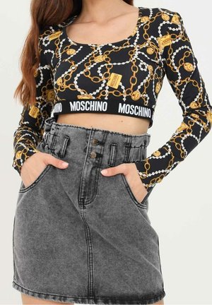 Kobieta ubrana w czarny krótki top z nadrukiem złotych łańcuchów i logo Moschino, w połączeniu z wysokim stanem szarej spódnicy dżinsowej, ręce w kieszeniach.