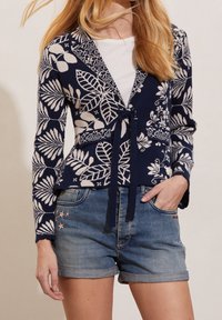 Námořnický cardigan s bílými květinovými vzory, véčkový výstřih, zavázaný v přední části, spárovaný se světle modrými džínovými šortkami s výšivkou hvězd a ohrnutými lemy.