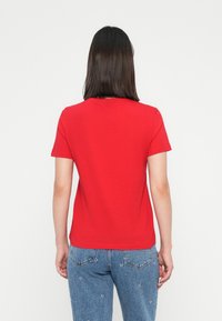 Woman with shoulder-length dark hair wearing a plain red short-sleeve T-shirt and blue jeans with subtle white floral patterns, viewed from behind.