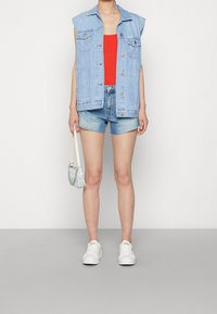 Denimväst med korta ärmar, ljusblå färg, lager på en röd tanktop. Bärs med blå shorts och vita sneakers, håller en liten handväska.