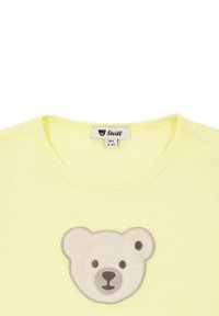 T-shirt di cotone giallo con applicazione di un orsetto di peluche in beige chiaro e marrone. Collo tondo e texture morbida. Etichetta del marchio Steiff visibile.