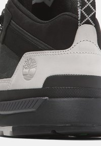 Černé a šedé tenisky se strukturovaným svrškem, hladkým bílým mezisolem a gumovou podešví se vzorovaným gripem. Logo vytlačené na boku.