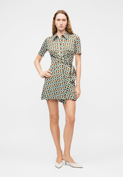 Νεαρή γυναίκα στέκεται με το χέρι στη μέση, φορώντας ένα φόρεμα με κοντά μανίκια και ζώνη, με γεωμετρικό μοτίβο και λευκά μυτερά τακούνια.