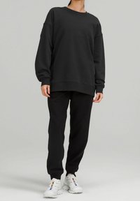 Felpa nera con spalle cadenti e maniche lunghe, abbinata a pantaloni jogger neri e scarpe da ginnastica bianche. Texture liscia, vestibilità comoda.