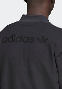 Svart sweatshirt med texturerat tyg och vikbar krage. "adidas"-logotypen och trefoten tryckt i mörka toner på baksidan.